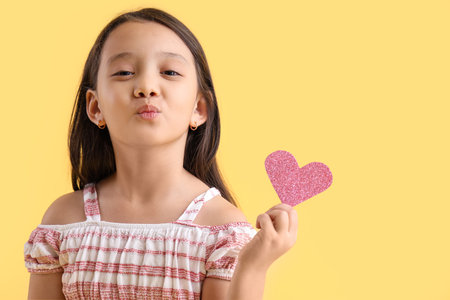 Cute little Asian girl with heart blowing kiss on yellow background. Valentine's Day celebrationの写真素材