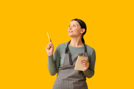Beautiful young waitress with notebook and pencil on yellow backgroundの写真素材