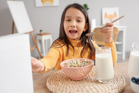 Cute little Asian girl eating cereal rings with milk at table in kitchenの写真素材