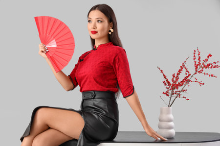 Young Asian woman with fan and berry branches on table against light background. Chinese New Year celebrationの写真素材