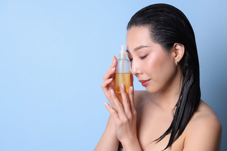Beautiful young Asian woman with bottle of hair oil on blue background, closeup