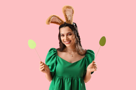 Beautiful woman in bunny ears with paper Easter eggs on pink background