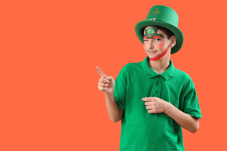 Funny boy with face painting and leprechaun's hat pointing at something on orange background. St....