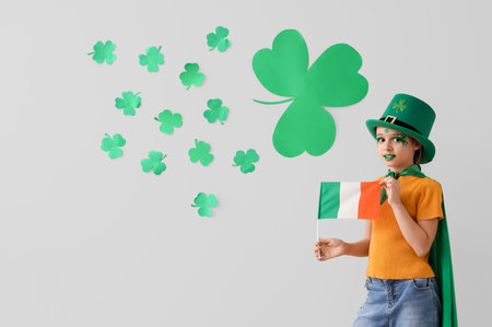 Funny girl with face painting, leprechaun's hat, flag of Ireland and clovers near white wall. St....
