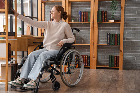 Young redhead woman in wheelchair taking book from shelf at libraryの写真素材