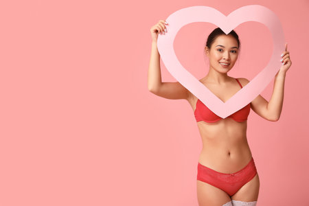 Beautiful young happy Asian woman in sexy underwear with heart-shaped frame on pink background. Valentine's Day celebrationの写真素材