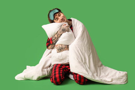 Sleepy young man with blanket, pillow and sleeping mask on green backgroundの写真素材