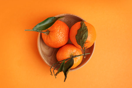 Wooden bowl with tasty tangerines and leaves on orange background