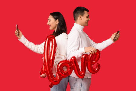 Young couple with mobile phones and air balloon in the shape of the word LOVE on red background. Online datingの写真素材