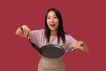 Portrait of young Asian woman with frying pan and spatula on red backgroundの写真素材