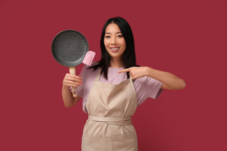 Portrait of young Asian woman pointing at frying pan and spatula on red backgroundの写真素材