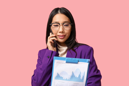 Portrait of young Asian businesswoman with clipboard talking by phone on pink backgroundの写真素材