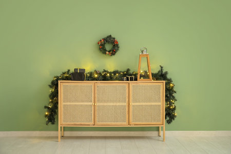 Dresser decorated for Christmas with garland, wreath and fir branches near green wall
