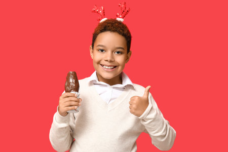 Cute little African-American boy in reindeer horns showing thumb-up gesture with chocolate Santa...