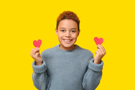 Cute little African-American boy with red paper hearts on yellow background. Valentine's day...