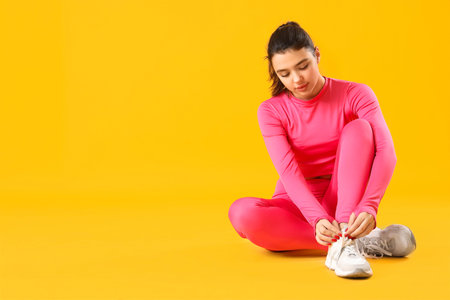Young woman in sportswear tying shoe laces on yellow backgroundの写真素材