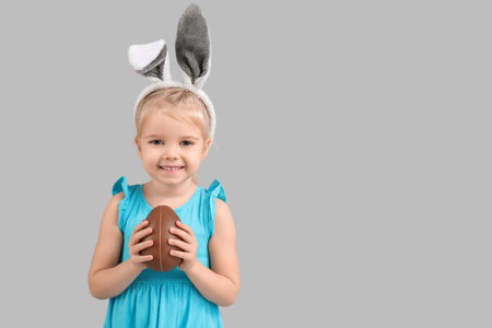 Happy little girl in bunny ears with chocolate egg on gray background. Easter celebration
