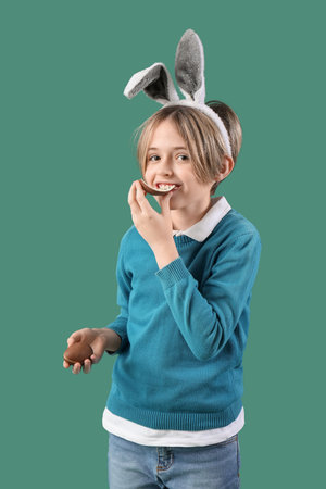 Cute little boy in bunny ears eating chocolate egg on green background. Easter celebration