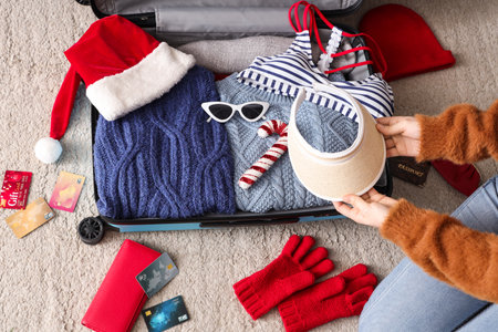 Woman packing beach accessories, clothes, credit cards and Christmas decor into suitcase at homeの写真素材