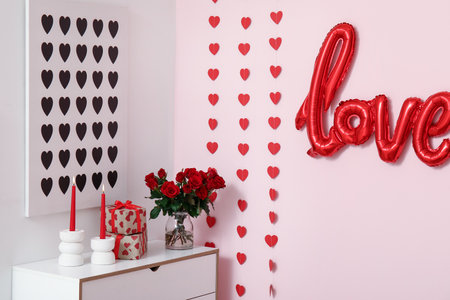 Bouquet of beautiful roses, candles and gift boxes on commode near pink wall in room decorated for Valentine's Day celebrationの写真素材