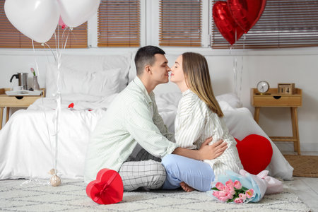 Beautiful young couple with gift box and bouquet of flowers sitting on the floor and celebrating Valentine's Day at homeの写真素材