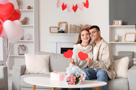 Beautiful young couple with gift box, bouquet of flowers and paper hearts celebrating Valentine's Day at homeの写真素材