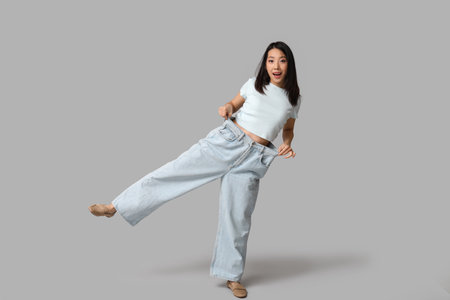 Beautiful young shocked Asian woman in loose jeans on gray backgroundの写真素材