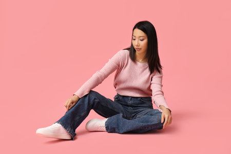 Beautiful young Asian woman in stylish jeans sitting on pink backgroundの写真素材