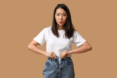Beautiful young displeased Asian woman buttoning up stylish jeans on brown backgroundの写真素材
