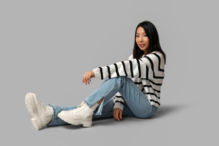 Beautiful young Asian woman in stylish jeans sitting on gray backgroundの写真素材