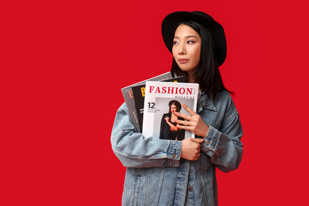 Beautiful young Asian woman in stylish denim jacket with magazine on red backgroundの写真素材