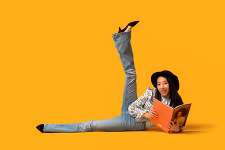 Beautiful young happy Asian woman in stylish jeans with magazine sitting on yellow backgroundの写真素材