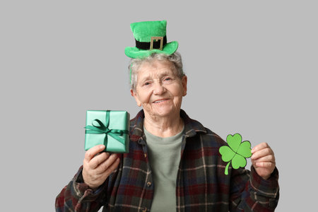 Senior woman in leprechaun hat with clover and gift box on gray background. St. Patrick's Day celebrationの写真素材