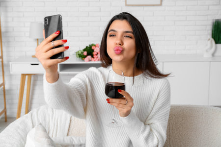 Young woman with glass of wine and mobile phone having online date at homeの写真素材