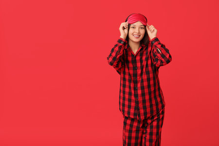 Beautiful young happy Asian woman in checkered pajamas with sleeping mask on red backgroundの写真素材