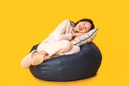 Beautiful young Asian woman in pajamas with pillow sleeping on beanbag chair against yellow backgroundの写真素材