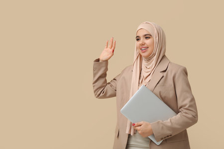 Beautiful young Muslim woman in hijab with modern laptop waving hand on beige backgroundの写真素材
