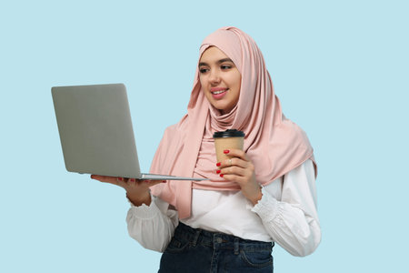 Beautiful young Muslim woman in hijab with paper cup of hot coffee and modern laptop on blue...