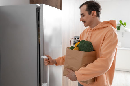 Young man with paper bag of fresh vegetables opening refrigerator in kitchenの写真素材