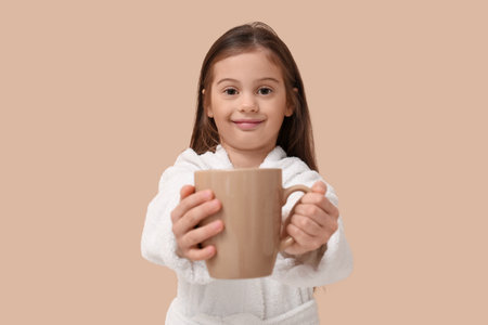 Cute little happy girl in bathrobe with cup of tea on beige backgroundの写真素材