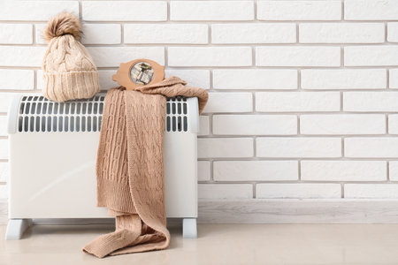 Electric radiator with piggy bank, hat and scarf near white brick wall. Heating saving conceptの写真素材