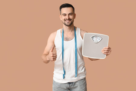 Handsome sporty young man with scales and measuring tape showing thumb-up gesture on brown background. Weight loss conceptの写真素材