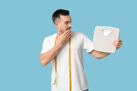 Handsome sporty shocked man with measuring tape and scales on blue background. Weight loss conceptの写真素材