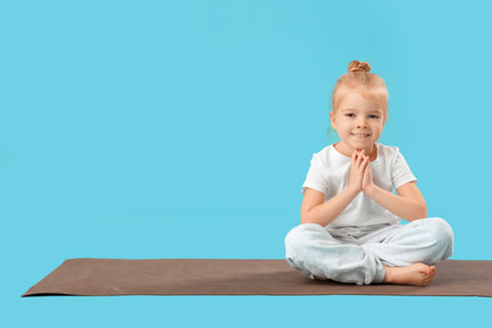 Cute little girl sitting in lotus pose on yoga mat against blue backgroundの写真素材
