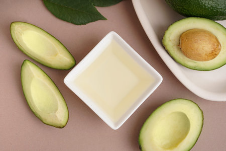 Bowl with essential oil and fresh ripe avocados on beige background