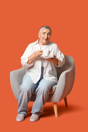 Senior woman with cup of tea sitting in armchair on orange backgroundの写真素材