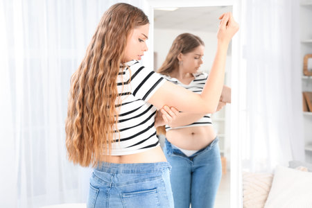 Young woman in tight jeans with chubby arms near mirror at home. Weight gain conceptの写真素材