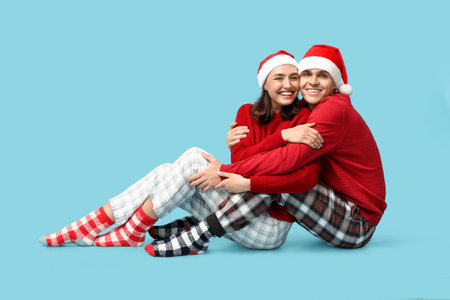 Happy young couple in Christmas pajamas and Santa hats sitting against blue backgroundの写真素材
