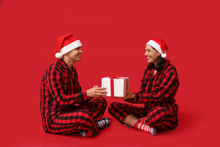 Happy young couple in Christmas pajamas and with gift on red backgroundの写真素材