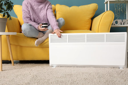 Young woman in warm winter clothes with cup of tea near electric heater. Concept of heating seasonの写真素材